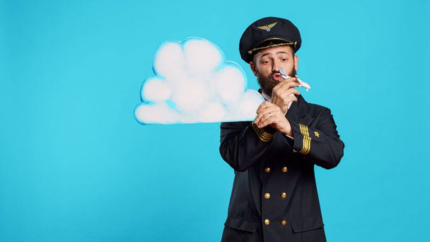 Male aviator playing with airplane toy and cloud board, holding cardboard icon and small miniature aircraft on camera. Young flying pilot with aviation uniform advertising aerial transport. - Foto, Imagem