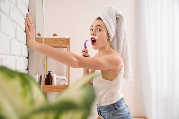 Молодая женщина бреется лицом с бритвой возле зеркала в ванной комнате - Фото, изображение