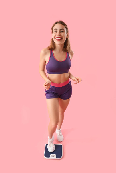 Sporty young woman measuring her weight on scales against pink background - Valokuva, kuva