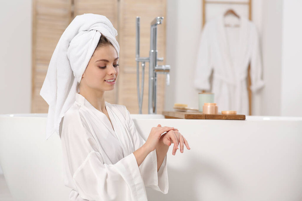 Beautiful woman in white robe applying cream near tub in bathroom - Foto, Bild
