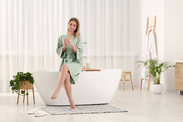 Beautiful happy woman in stylish robe with cup of hot drink sitting on tub in bathroom - Zdjęcie, obraz