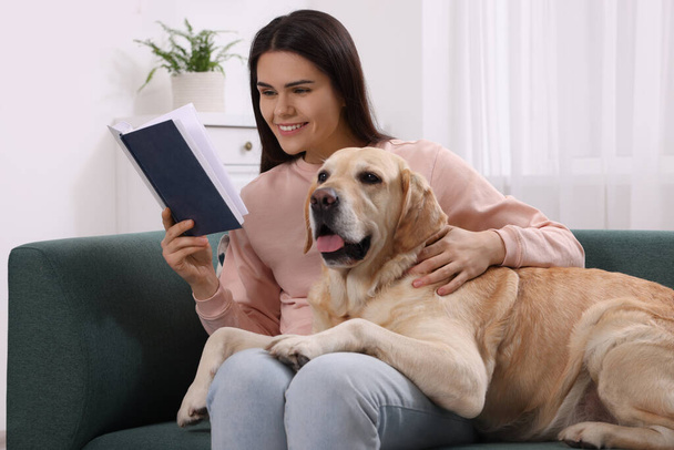 Happy woman reading book while sitting with cute Labrador Retriever on sofa at home - Foto, imagen