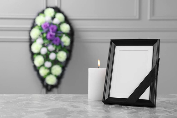 Photo frame with black ribbon, burning candle on light grey table and wreath of plastic flowers near wall indoors, space for text. Funeral attributes - Zdjęcie, obraz