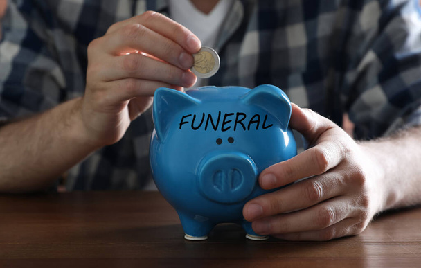 Money for funeral expenses. Man putting coin into piggy bank at wooden table, closeup - Фото, изображение
