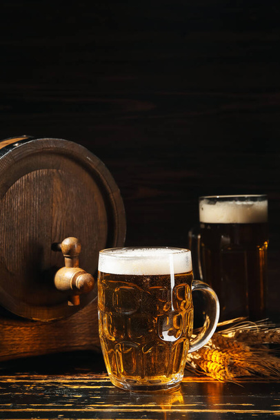 Becher mit kaltem Bier und Holzfass auf dunklem Hintergrund - Foto, Bild