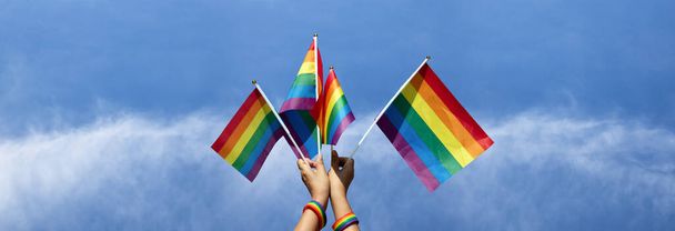 Drapeaux arc-en-ciel tenant dans les mains sur fond nuageux et bleu, concept pour appeler toutes les personnes à soutenir les personnes LGBT et à respecter la diversité des genres des gens à travers le monde. - Photo, image