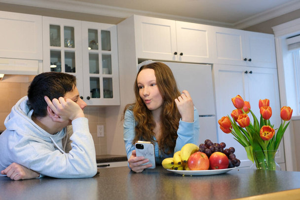 comunicazione in cucina amici Odnoklassniki Mangiare uva frutta in possesso di un telefono in mano una ragazza adolescente e un ragazzo sullo sfondo di armadi da cucina bianchi fiori tulipani in tuta - Foto, immagini