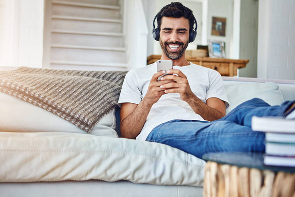 Músicas incríveis para o tempo solo. um homem relaxando no sofá enquanto usa seu telefone e fones de ouvido - Foto, Imagem
