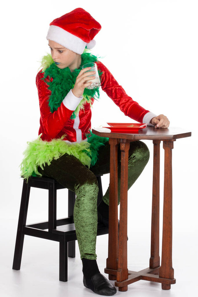 Vollständiges Porträt eines jungen Mädchens mit rotem Weihnachtsmannhut wie ein Grinch, das mit Milch in der Hand, isoliert auf weißem Hintergrund, hinter sich glüht. Konzept für Elfen - Foto, Bild