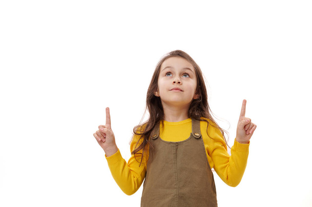 Petite fille blanche 5-6 ans regardant de côté, pointant ses index vers le haut sur l'espace publicitaire de copie sur fond blanc isolé. Portrait d'enfant intelligent intelligent préscolaire, écolière - Photo, image