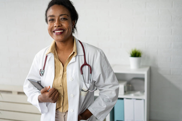 femme médecin afro-américaine souriante avec stéthoscope et manteau blanc debout à l'hôpital  - Photo, image