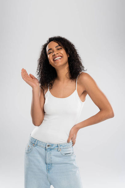 joyful african american woman in white tank top waving hand and smiling isolated on grey - Fotoğraf, Görsel