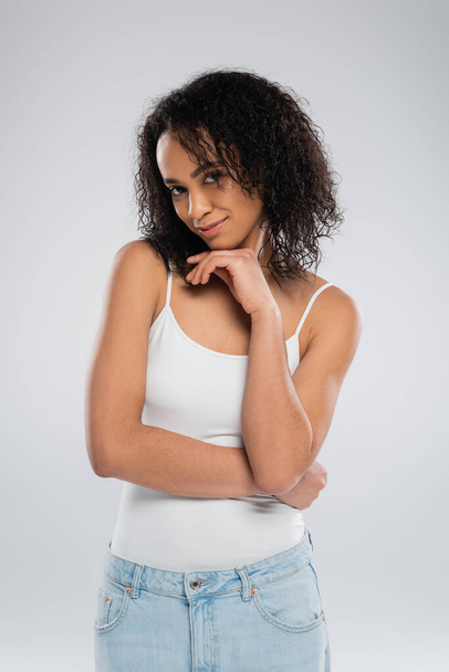flirty and smiling african american woman in white tank top holding hand near face and looking at camera isolated on grey - Fotó, kép