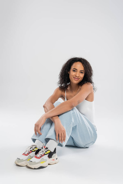 full length of african american woman in blue jeans and sneakers sitting and smiling at camera on grey background - Foto, immagini