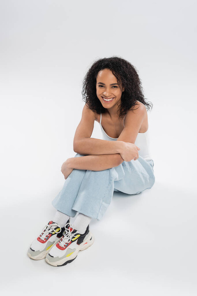 full length of cheerful african american woman in sneakers and jeans sitting on grey background - Φωτογραφία, εικόνα