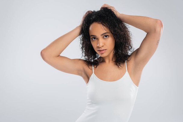young african american woman in white tank top touching wavy brunette hair and looking at camera isolated on grey - Fotografie, Obrázek