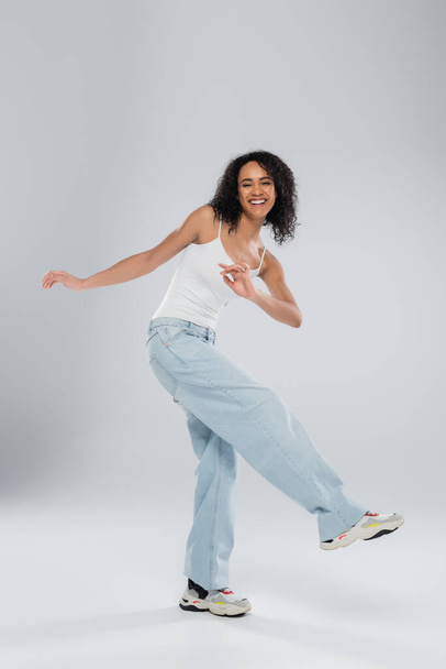 full length of carefree african american woman in white tank top and jeans posing on grey background - Foto, Bild