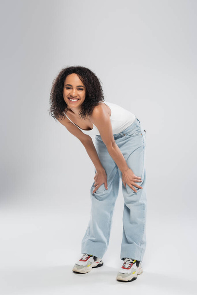 full length of carefree african american woman in blue jeans smiling at camera on grey background - Foto, immagini