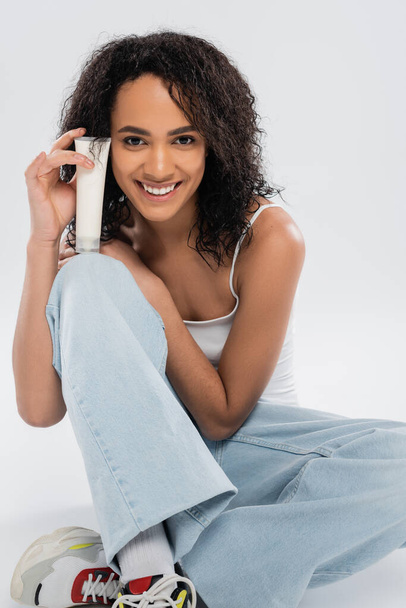 smiling african american woman holding tube of cosmetic cream while sitting on grey background - Fotó, kép