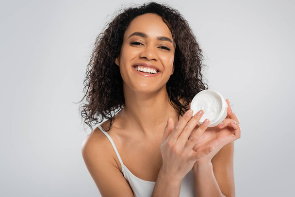 joven y feliz mujer afroamericana con piel perfecta tocando crema cosmética aislada en gris - Foto, imagen