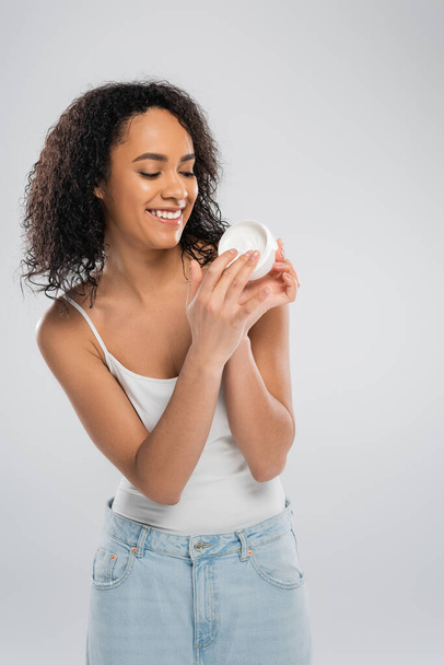 pleased african american woman in tank top looking at container of cosmetic cream isolated on grey - Foto, imagen
