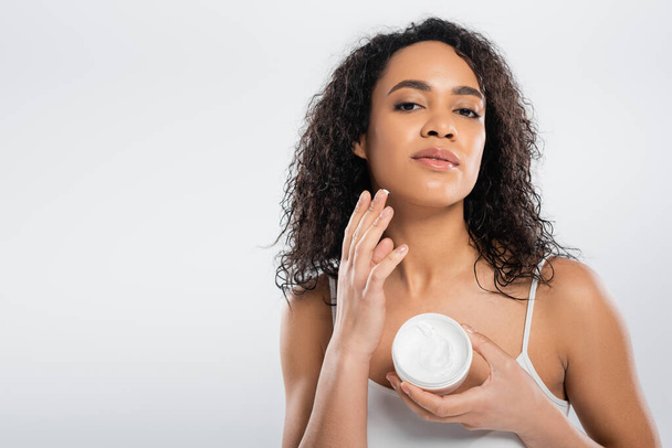 charming african american woman applying face cream while looking at camera isolated on grey - Foto, afbeelding