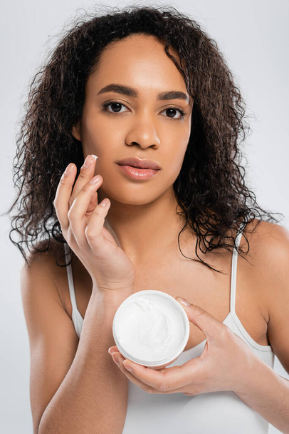 african american woman with curly brunette hair applying face cream isolated on grey - Foto, afbeelding
