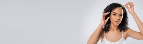brunette african american woman in white tank top applying cosmetic serum isolated on grey, banner - Foto, Imagem