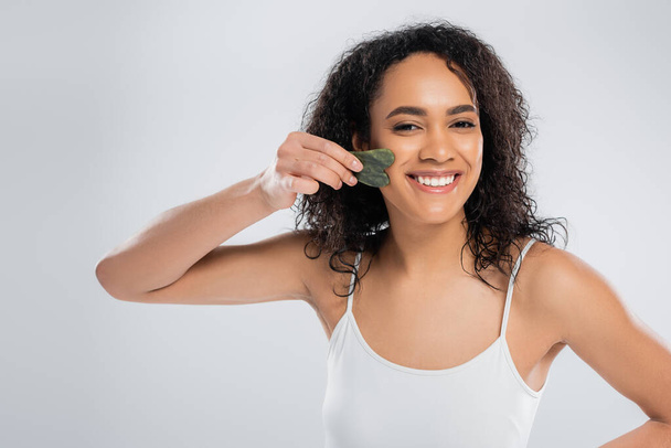 happy african american woman doing face massage with jade scraper isolated on grey - Photo, Image