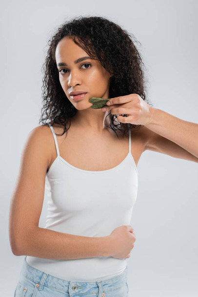 young african american woman in white tank top using face scraper isolated on grey - Foto, Bild
