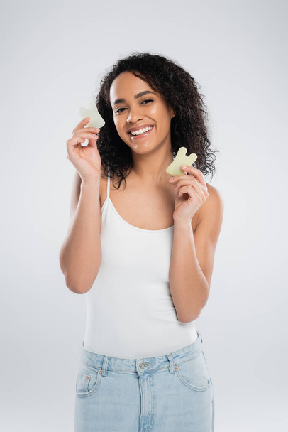 брюнетка африканская американка держит каменные скребки и улыбается в камеру, изолированную на сером - Фото, изображение