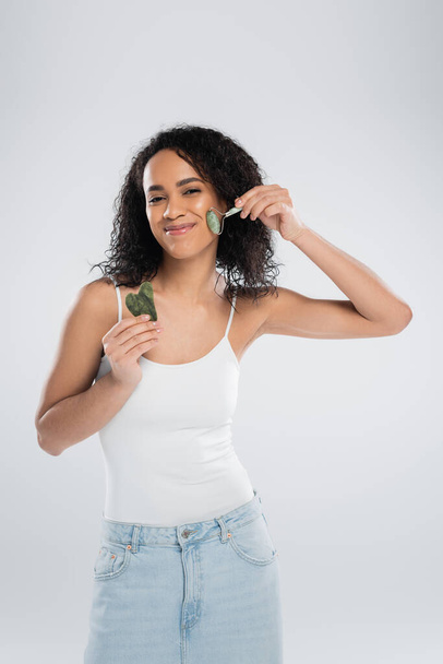 zadowolona african amerykański kobieta trzyma skrobak twarz i za pomocą jade roller izolowane na szary - Zdjęcie, obraz