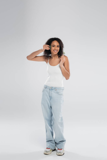 longitud completa de la sonriente mujer afroamericana en camiseta sin mangas y jeans posando con exfoliante facial y rodillo de jade sobre fondo gris - Foto, imagen