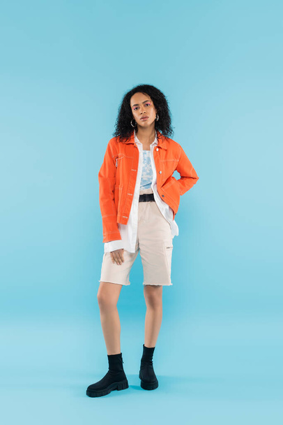 full length of african american woman in bright orange jacket and cotton shorts posing with hand on hip on blue background - Φωτογραφία, εικόνα