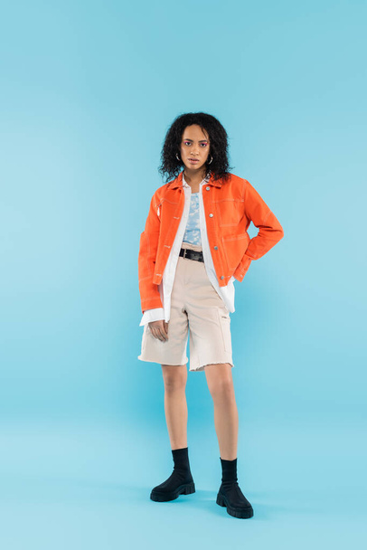 full length of african american woman in cotton shorts and orange jacket posing with hand on hip on blue background - Fotografie, Obrázek
