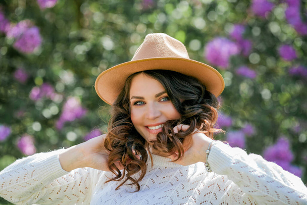 Portrait of beautiful young dark-haired woman with blue eyes in hat near the blooming. Happy model with curly hair. - Foto, Imagen