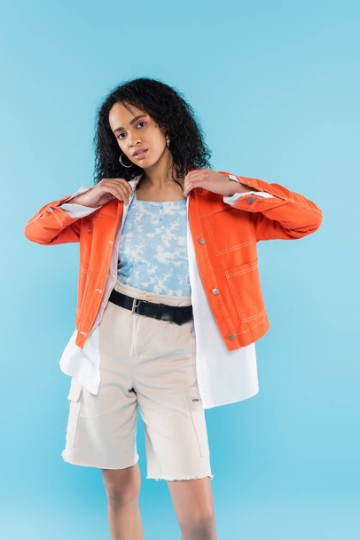 curly african american woman in white shorts adjusting bright orange jacket isolated on blue - Фото, изображение