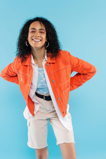 cheerful african american woman in stylish orange jacket smiling at camera isolated on blue - Foto, imagen