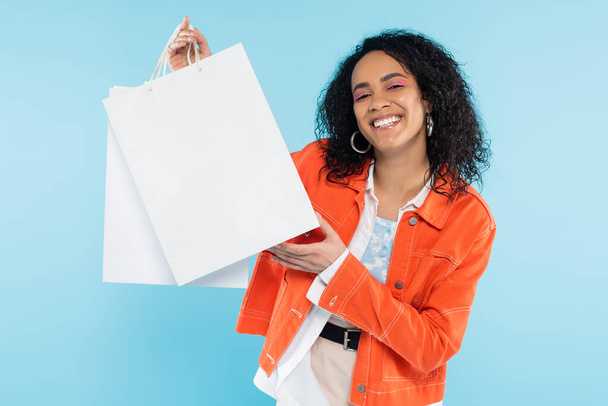boldog afro-amerikai nő narancssárga dzsekiben kezében fehér bevásárló táskák elszigetelt kék - Fotó, kép