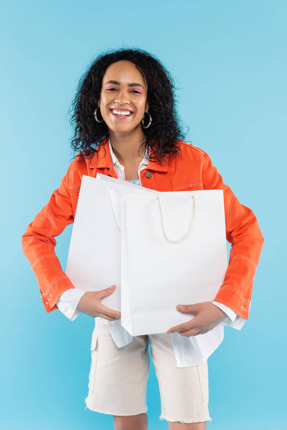módní africká americká žena s bílými nákupními taškami s úsměvem na kameru izolované na modré - Fotografie, Obrázek