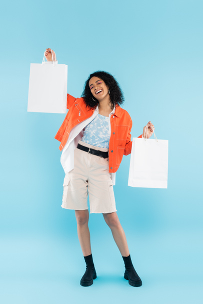 full length of cheerful and trendy african american woman posing with white shopping bags on blue background - Foto, afbeelding
