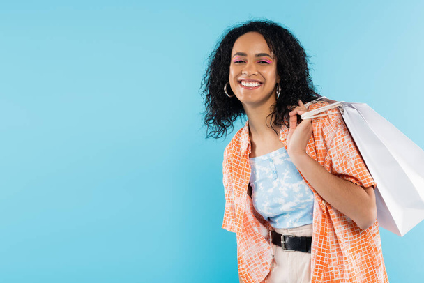 elegante donna africana americana con capelli ricci bruna che tiene borse della spesa e sorridente alla fotocamera isolata sul blu - Foto, immagini