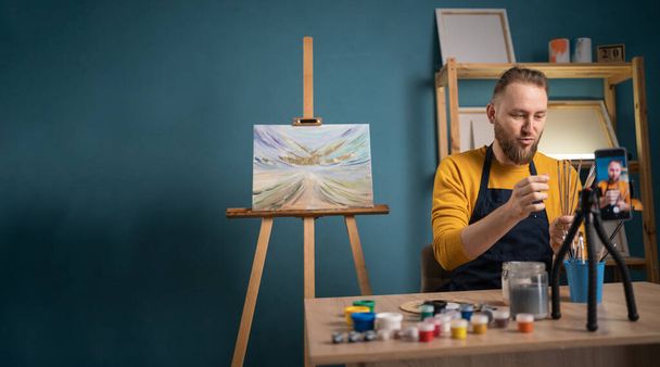 Fiatal művész férfi ül íróasztal és rögzíti az online művészeti osztály a művészeti stúdióban. Blogger csinál egy videót a blogja művészet mutatja akvarell festék. Másold a helyet. Zászlós - Fotó, kép