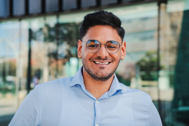 close up portrait of hispanic young entrepreneur smiling and looking at camera at workspace. Front view of one latin happy employee office worker with glasses and successful attitude standing outdoors - Photo, image