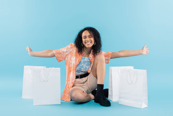 comprimento total de mulher americana africana alegre sentado com as mãos estendidas perto de sacos de compras brancos no fundo azul - Foto, Imagem