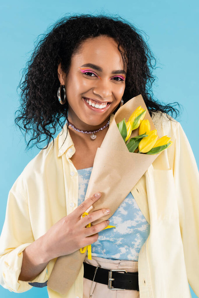 happy and stylish african american woman holding yellow tulips isolated on blue - Foto, afbeelding