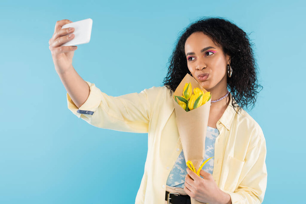brunette african american woman with yellow tulips pouting lips and taking selfie on cellphone isolated on blue - 写真・画像