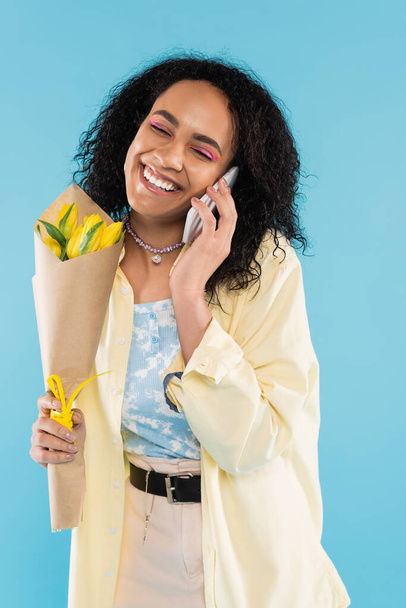 pleased african american woman with yellow tulips and closed eyes talking on mobile phone isolated on blue - Foto, imagen