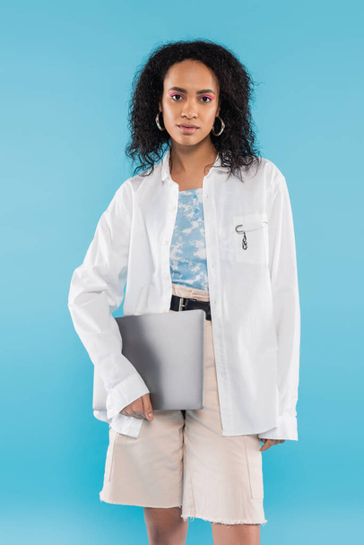 brunette african american woman in white shirt holding laptop and looking at camera isolated on blue - Photo, Image
