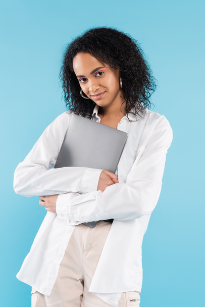 happy african american woman in white shirt standing with laptop and looking at camera isolated on blue - Foto, Imagem